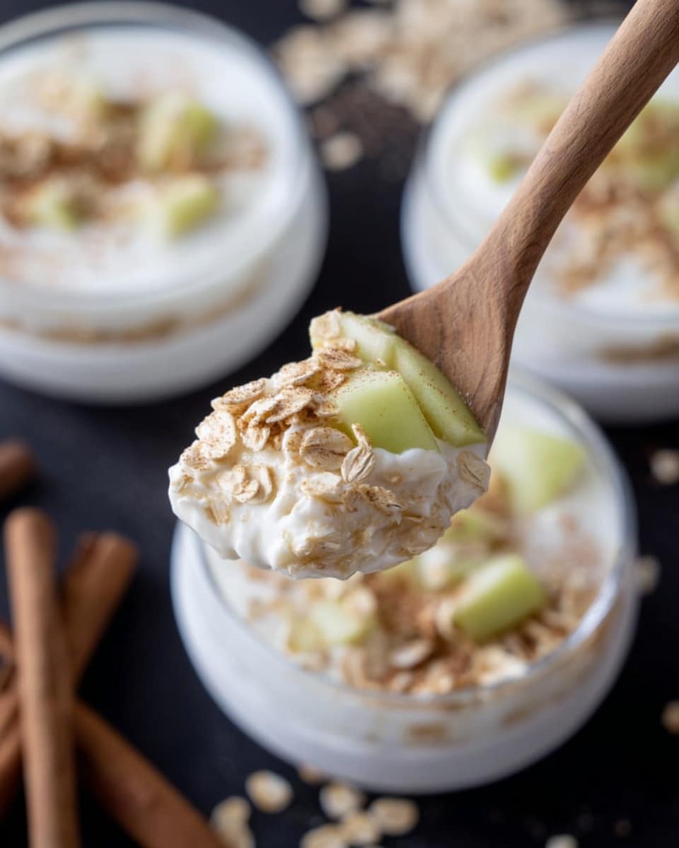 A close-up view of a wooden spoon holding a scoop of a layered dish in a white bowl. The dish has three clear layers: the bottom layer is white yogurt, the middle layer consists of small, light green apple pieces, and the top layer is a sprinkle of light beige rolled oats. In the background, two more white bowls filled with the same dish are softly focused, placed on a white marbled surface. Scattered rolled oats and cinnamon sticks add to the setting. Photo taken with an iphone --ar 4:5 --v 7
