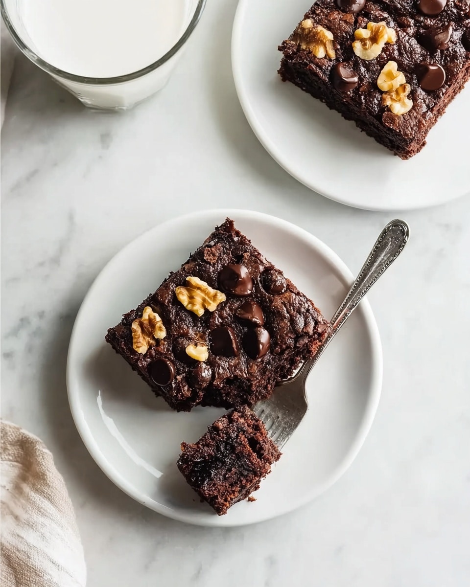 A square slice of rich, dark brown chocolate brownie sits on a white plate, topped with visible chunks of walnuts and glossy chocolate chips. The texture of the brownie appears moist and slightly crumbly, with a bite taken out and held on a silver fork resting on the plate. Nearby, another similar slice is on a second white plate, with a similar dark rich color and nut and chocolate chip toppings. To the left, a glass of milk is partially visible. All items are placed on a white marbled surface. Photo taken with an iphone --ar 4:5 --v 7