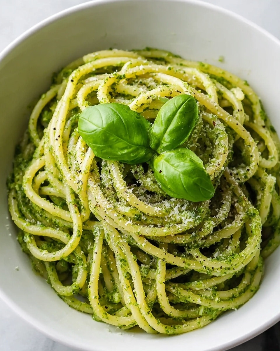 A close-up view of a white bowl filled with spaghetti pasta coated evenly with green pesto sauce, showing a slightly glossy texture. The pasta noodles are thick and curled, twirled neatly in the bowl, with a sprinkle of grated cheese and black pepper lightly dusted across the top. At the center, two bright green fresh basil leaves rest on the pasta as garnish. The bowl sits on a white marbled surface, adding a clean and simple background to highlight the vibrant colors of the dish. Photo taken with an iphone --ar 4:5 --v 7