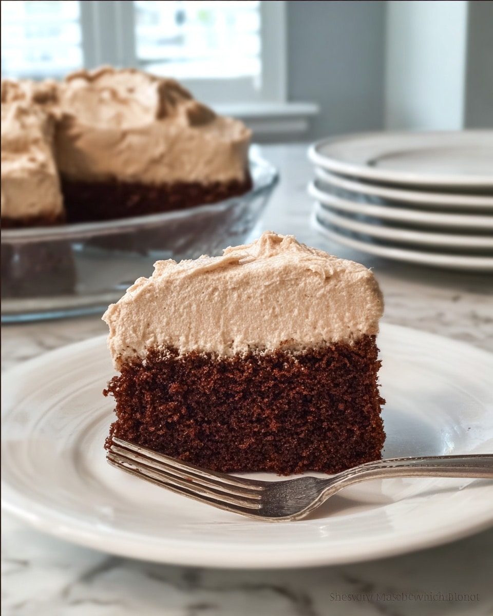A single slice of two-layer cake sits on a white plate with a silver fork resting beside it. The bottom layer is a thick, dark brown, moist cake with a slightly rough texture. On top, there is a generous layer of light brown frosting with a whipped, fluffy texture. In the background, the rest of the cake is visible inside a clear glass round dish. The scene is set on a white marbled surface with blurred windows and stacked white plates in the back. photo taken with an iphone --ar 4:5 --v 7