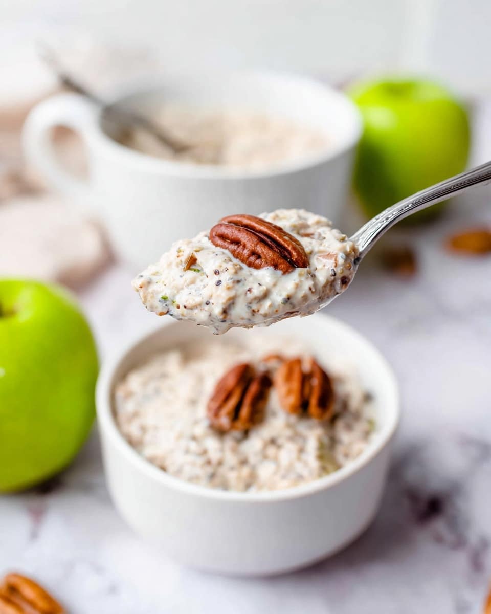 A close-up of a silver spoon holding a creamy mixture with small visible seeds and bits, hovering over a white bowl containing more of the same mixture topped with three pecan halves. The mixture is light beige with brown specks, with a thick and slightly chunky texture. In the background, there are two whole green apples and another white bowl with the same mixture, all placed on a white marbled surface. The overall setting is bright with soft natural lighting. photo taken with an iphone --ar 4:5 --v 7