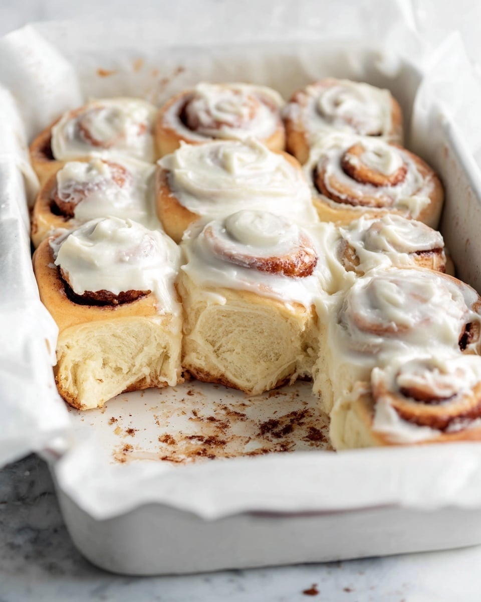 A white rectangular baking dish filled with nine cinnamon rolls arranged in three rows and three columns. Each cinnamon roll has a golden-brown spiral base topped with a thick, creamy white frosting that slightly melts down the sides. The rolls are touching each other and are lined with white parchment paper that folds over the edges of the dish. The dish sits on a white marbled surface with a white cloth napkin partly under it, and a small stack of white plates is visible in the upper right corner. Photo taken with an iphone --ar 4:5 --v 7
