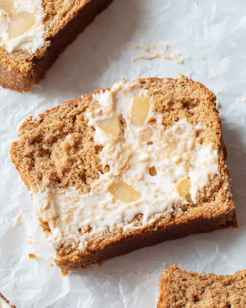 The image shows a close-up of a single slice of bread with a rough, crumbly texture and a light brown color. The slice is spread with a white, creamy layer that appears to be melting or glistening, covering parts of the surface unevenly. Small chunks of a pale yellow ingredient, likely apple pieces, are embedded within the bread, visible mostly towards the middle. The edges of the slice are darker brown and slightly crisp. The slice lies on a white marbled textured surface with another partially visible bread piece in the background. photo taken with an iphone --ar 4:5 --v 7