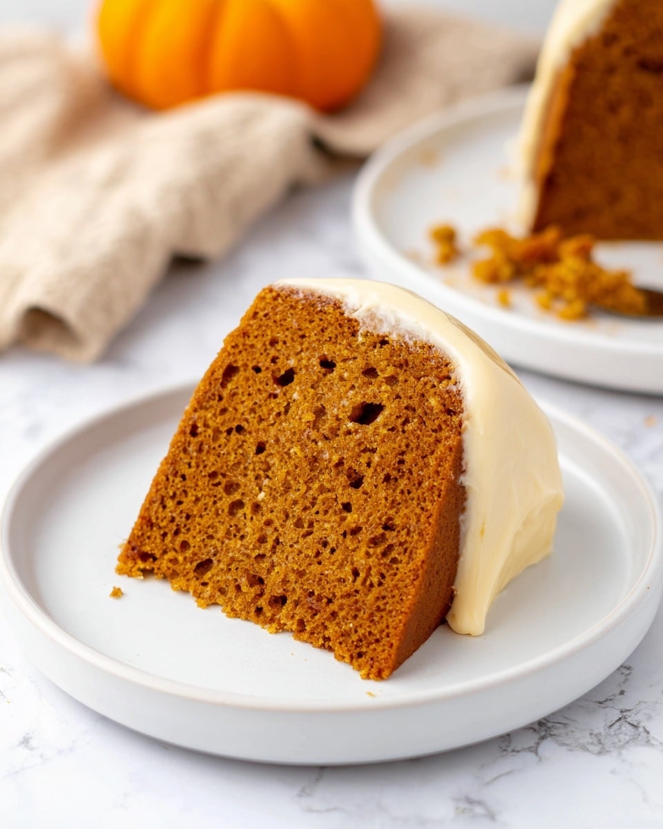 A single slice of moist pumpkin cake with a deep orange-brown color sits on a simple white round plate. The cake has one visible thick layer with a dense texture and small air holes throughout. At the base of the slice is a smooth layer of pale cream-colored frosting that wraps around the bottom edge. In the background, there is another slice on a white plate with some crumbs scattered nearby, and a small orange pumpkin sits on a beige cloth, all set on a white marbled surface. photo taken with an iphone --ar 4:5 --v 7