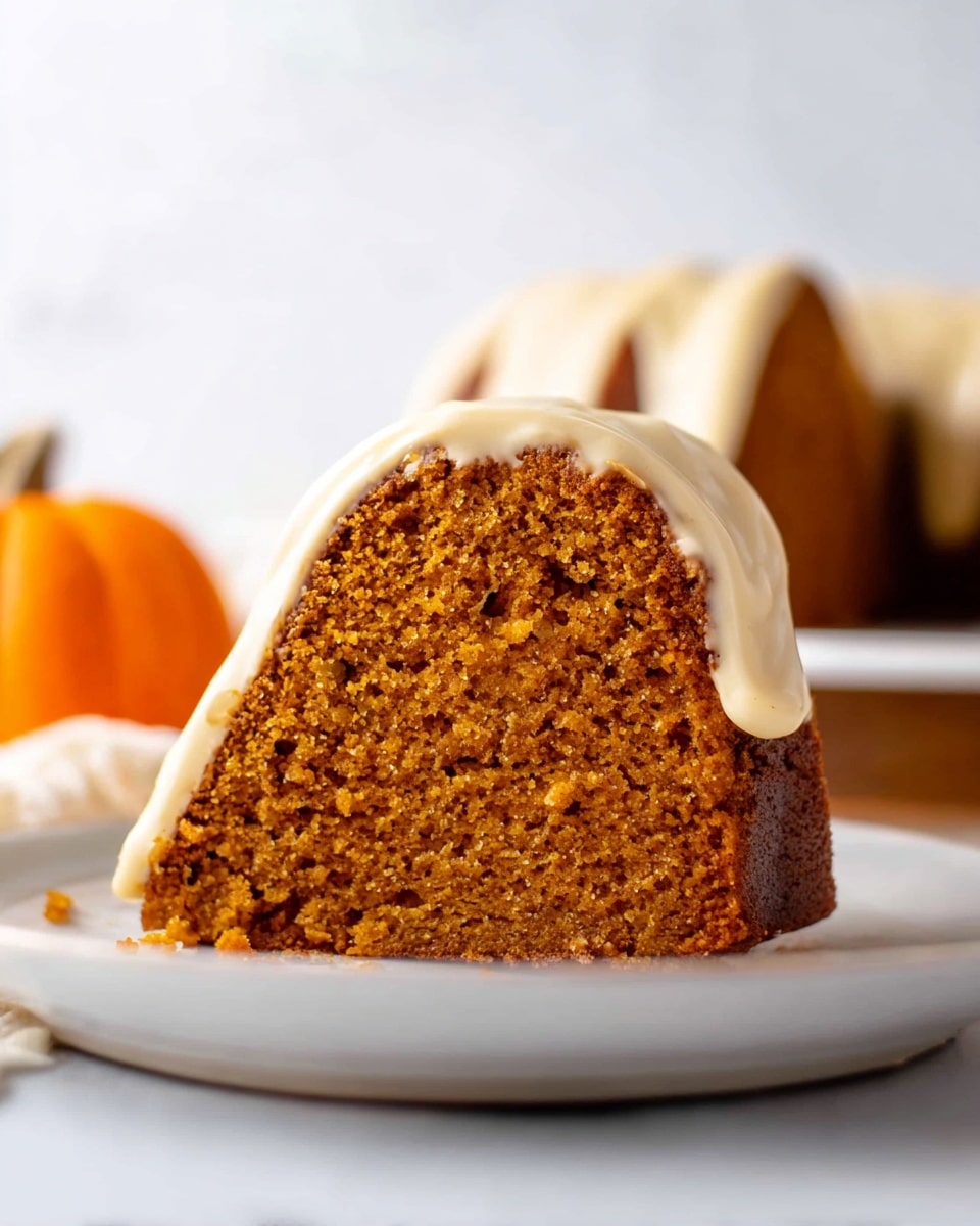 A slice of pumpkin bundt cake sits on a white plate with a close-up view. The cake has two main layers: the dense, moist, brownish-orange cake layer with a slightly crumbly texture forms the base, and a thick, creamy off-white glaze smoothly drapes over the top and sides of the cake. The background features a soft white marbled texture, with a small, faint orange pumpkin blurred in the distance, adding an autumn feel. Photo taken with an iphone --ar 4:5 --v 7