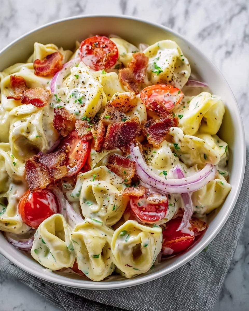 A large white bowl filled with creamy tortellini pasta as the base layer, with its smooth, folded shapes coated in white sauce speckled with green herbs. Mixed throughout are bright red cherry tomato halves and thin pale purple onion rings adding vibrant color and a slightly translucent texture. Crispy chunks of reddish-brown bacon are scattered over the top, providing a crunchy contrast. Yellow-green pepper slices are also visible among the layers, while a sprinkle of black pepper and herbs tops the dish. The bowl sits on a light gray cloth over a white marbled surface. photo taken with an iphone --ar 4:5 --v 7