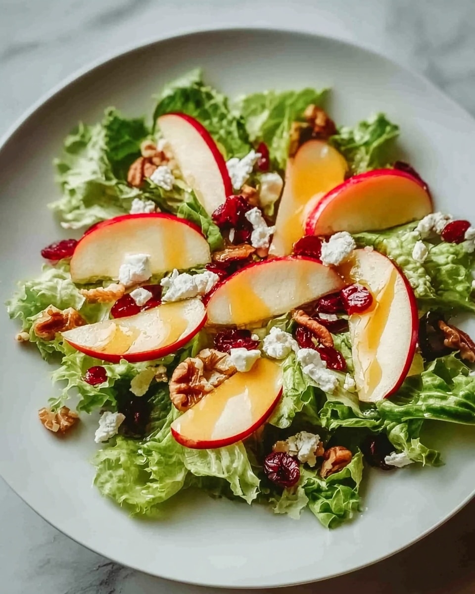 The image shows a fresh salad on a white plate set on a white marbled surface, featuring a base layer of bright green leafy lettuce. On top, there are thin slices of red-skinned apple arranged evenly across the lettuce. Scattered around are small chunks of creamy white cheese and reddish-brown nuts that add texture. The salad is drizzled with a glossy, light yellow dressing that adds a little shine. photo taken with an iphone --ar 4:5 --v 7