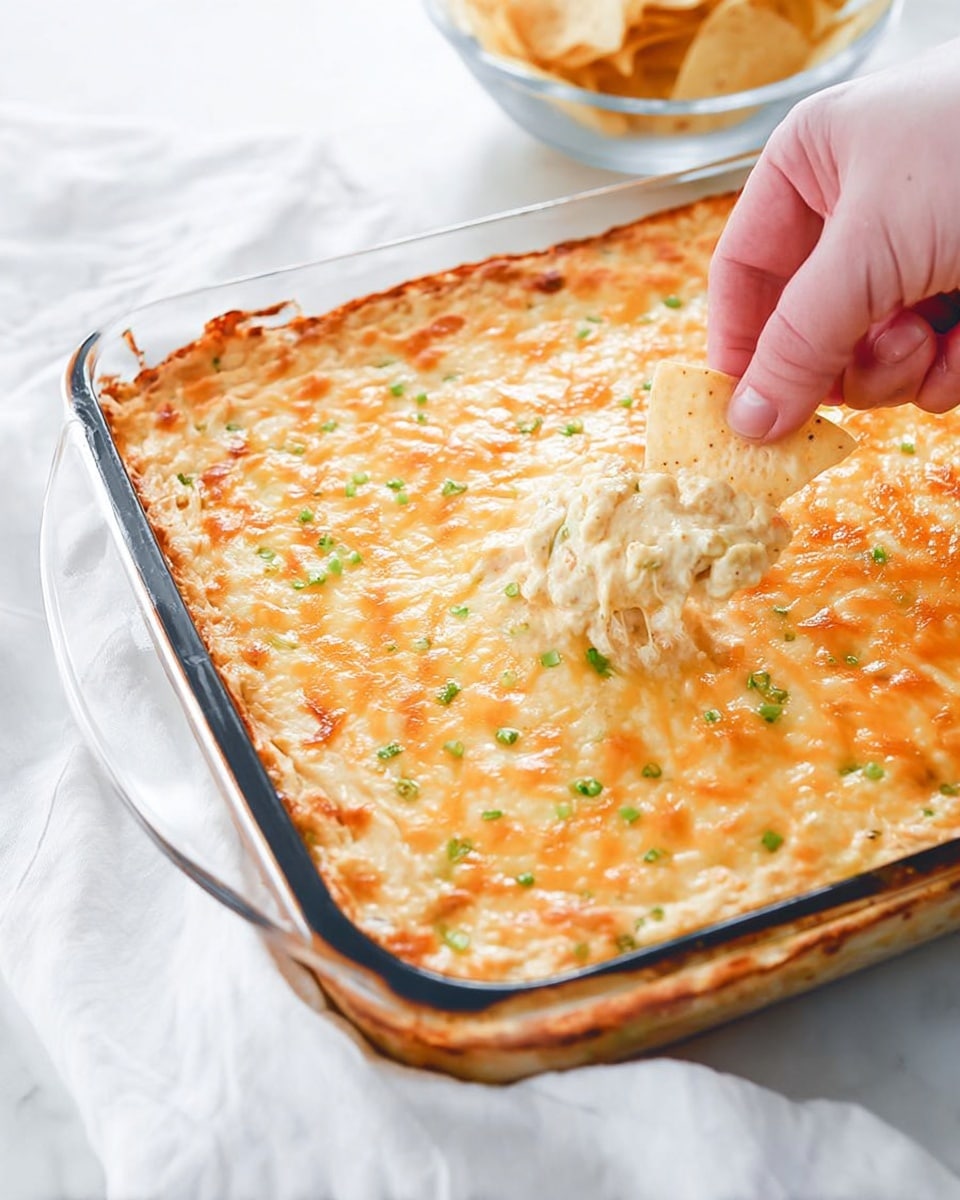 A glass baking dish filled with a creamy, cheesy dip that has a golden-brown melted cheese crust on top, sprinkled with small green bits of herbs evenly spread across the surface. Near the bottom right corner, a woman's hand is dipping a light tan, slightly crisp chip into the creamy dip beneath the melted cheese layer. The dish sits on a white marbled background with a white cloth underneath, and a clear bowl of more chips is partially visible in the background. The overall look is warm and inviting with a soft, gooey texture under the crust. photo taken with an iphone --ar 4:5 --v 7