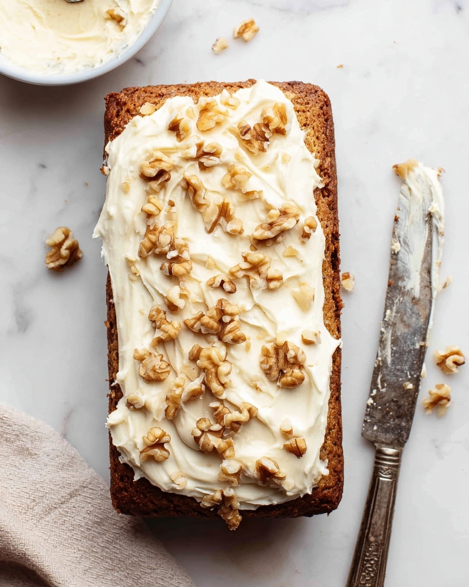 A rectangular loaf cake with a dark brown base layer topped with a thick, creamy white frosting spread unevenly across the top, showing soft, swirled texture. Scattered on the frosting are chopped light brown walnuts, adding rough texture and contrast. The cake is placed on a white marbled surface with a vintage knife beside it, showing some frosting residue. There is also a small white bowl with some more creamy frosting partially visible at the top left and a soft, beige cloth at the bottom left. Photo taken with an iphone --ar 4:5 --v 7