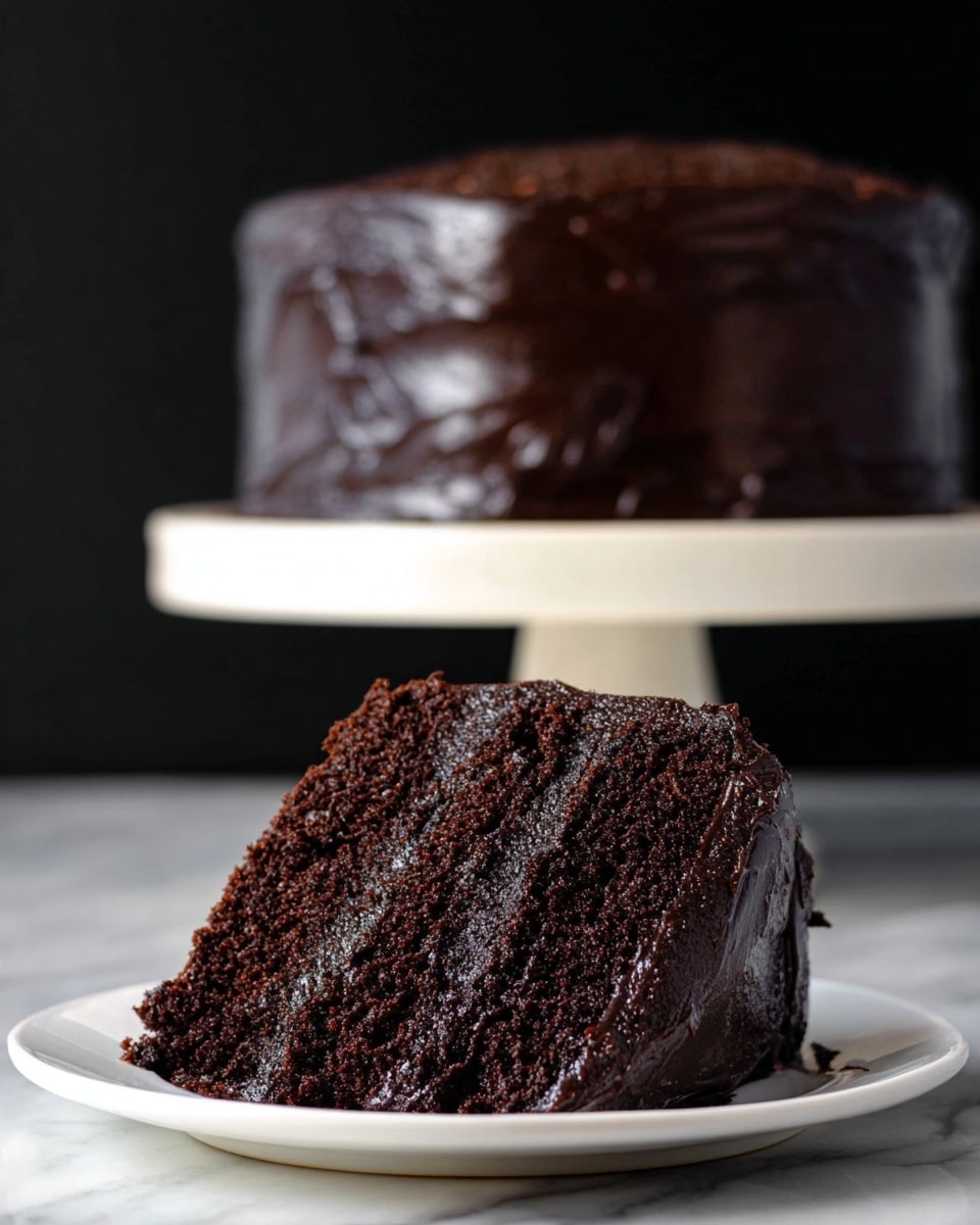 A thick slice of dark chocolate cake with two visible layers is placed on a white plate; the cake looks moist with a rich, dense crumb texture and is covered fully in smooth, glossy dark chocolate frosting that wraps around the sides and top, creating a shiny and thick layer. In the background, a whole cake same as the slice stands on a white cake stand, with the dark chocolate texture slightly blurred against a dark backdrop, all set on a white marbled surface. photo taken with an iphone --ar 4:5 --v 7