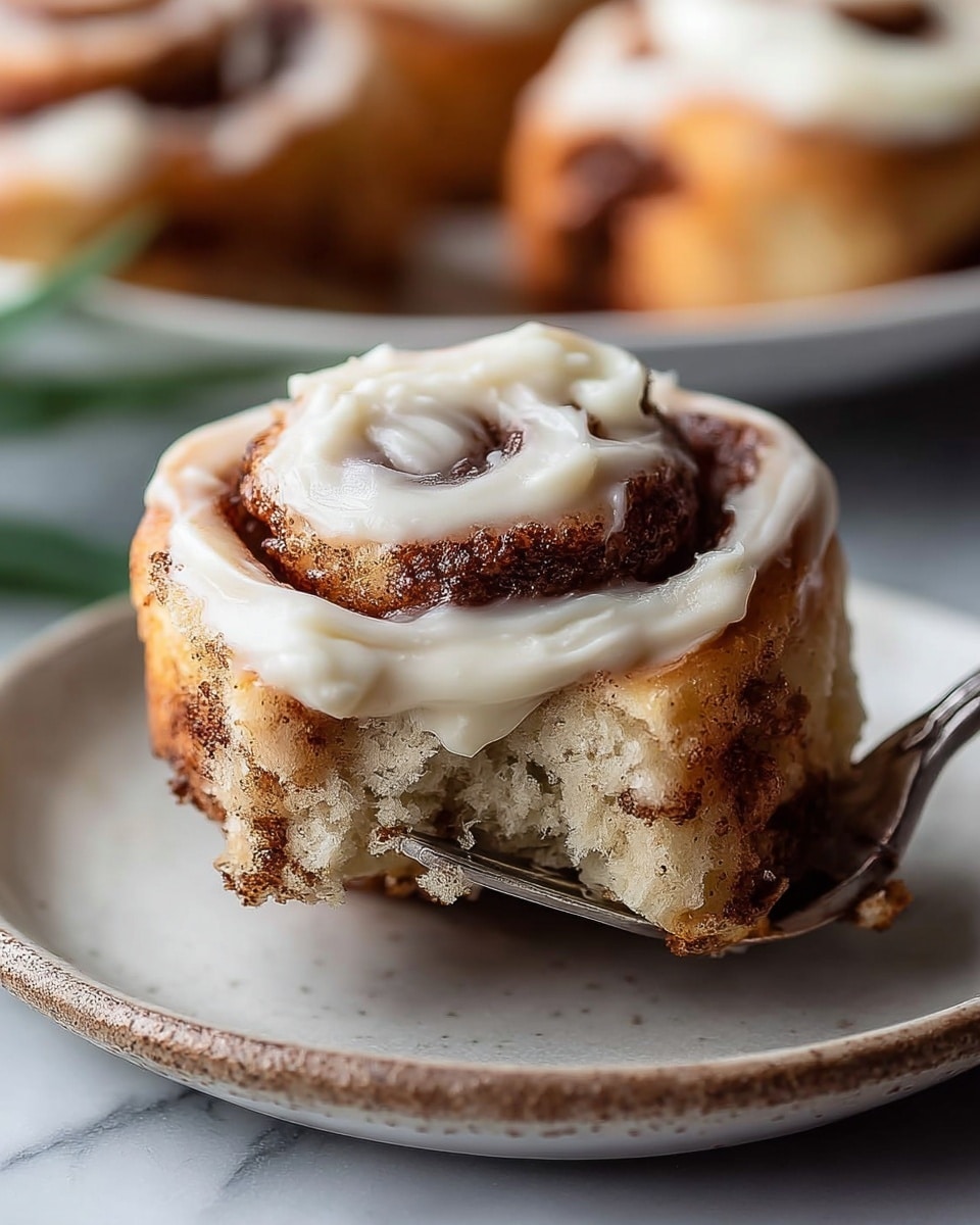 A close-up view of a single cinnamon roll with three visible layers: a golden brown base with a slightly crispy texture, a middle soft and fluffy dough layer sprinkled with dark brown cinnamon swirls, and the top covered with thick smooth white cream cheese frosting spread unevenly that highlights the spiral shape. The cinnamon roll is held by a silver fork lifting it from a white ceramic plate with a slightly rustic edge. The background is a soft-focus white marbled surface with blurred other cinnamon rolls visible. The lighting is natural and warm, enhancing the roll's moist and soft textures. Photo taken with an iphone --ar 4:5 --v 7