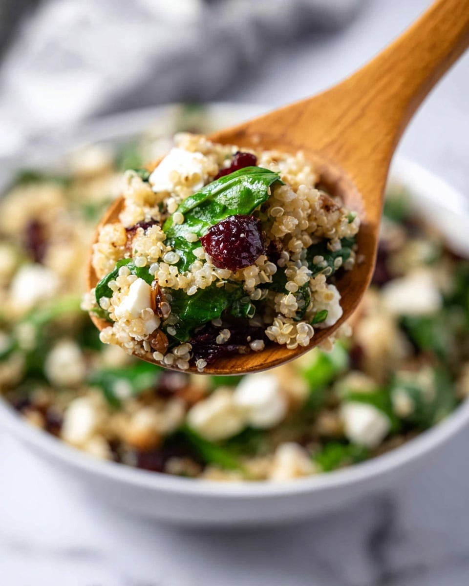 A close-up of a wooden spoon holding a colorful quinoa salad above a white bowl filled with the salad, all placed on a white marbled surface. The salad has small, round, light beige quinoa grains mixed with bright green fresh spinach leaves, cubed white cheese, and dark red dried cranberries. The background is softly blurred to keep the focus on the spoonful of salad. Photo taken with an iphone --ar 4:5 --v 7