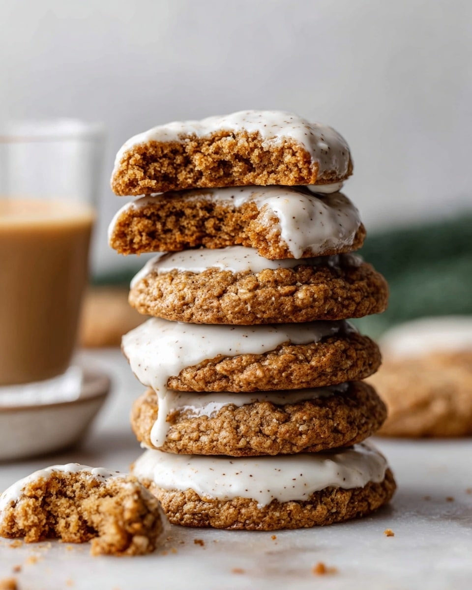 A stack of five oatmeal cookies with a rough texture and a golden-brown color is placed on a white marbled surface. Each cookie is partially dipped in white frosting that has small specks throughout, with the frosting mainly covering the lower half of each cookie, creating a contrast against the cookie’s rough surface. The top cookie is broken in half, showing the soft, crumbly interior with a slightly darker shade of brown. Crumbs are scattered near the base of the stack, with a small bit of cookie piece broken off sitting in the foreground on the left. The background features a blurred glass with a tan drink and a small white dish partially out of focus. photo taken with an iphone --ar 4:5 --v 7
