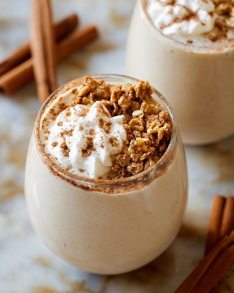 A creamy beige smoothie fills a clear rounded glass, topped with three distinct sections: one quarter is sprinkled with fine brown cinnamon powder, the middle quarter has dollops of white whipped cream with a smooth texture, and the last half is covered with crunchy light brown granola pieces, showing varied textures and small clusters. The glass sits on a white marbled surface with blurred cinnamon sticks lying around in the background. Another similar smoothie is partially visible in the upper right corner, showing the same topping pattern. photo taken with an iphone --ar 4:5 --v 7