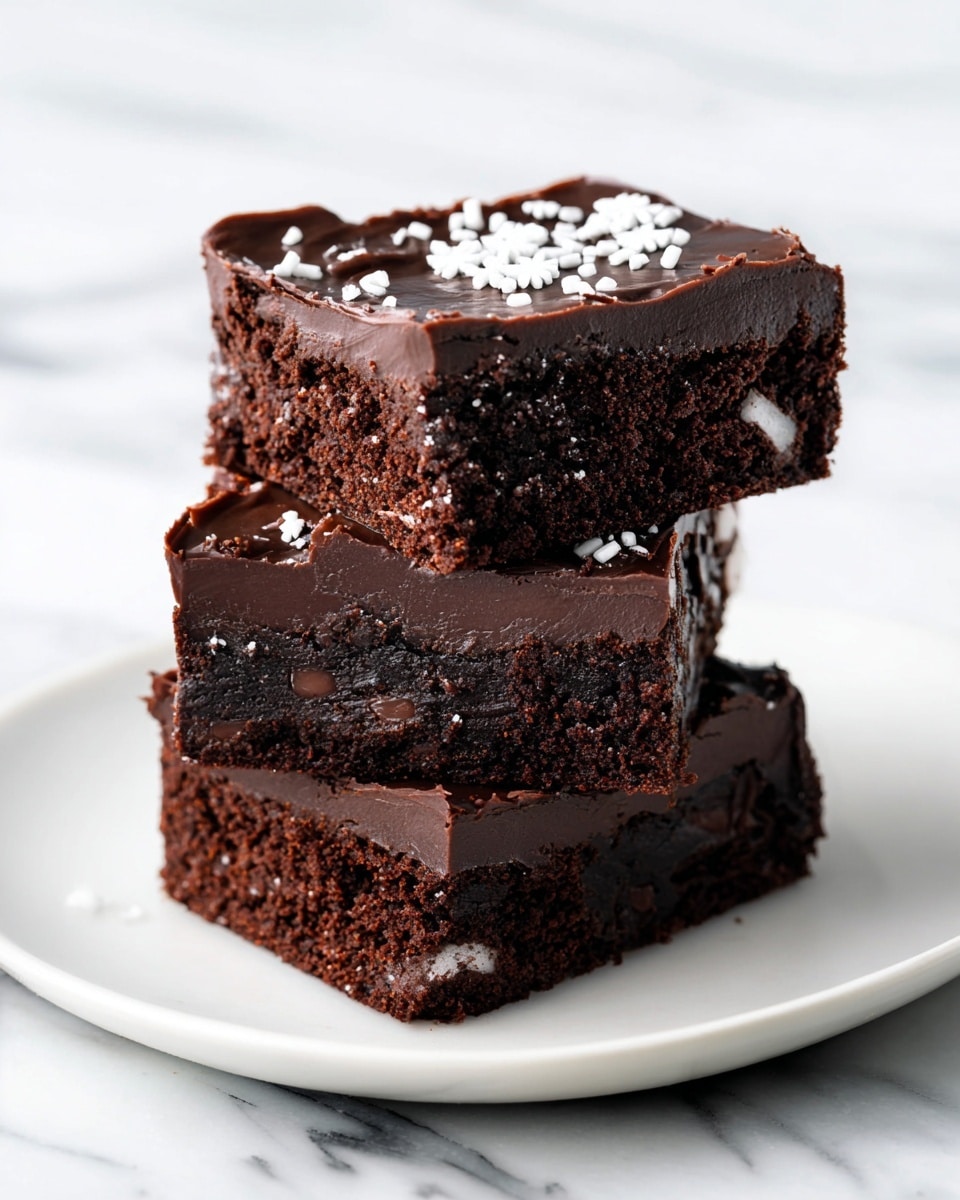 The image shows seven dark chocolate brownies arranged on a white round plate with a white marbled texture underneath. Each brownie is a square with two thick layers: a dense, moist dark brown base and a shiny, smooth darker brown chocolate frosting on top. The frosting is decorated with small white sprinkles shaped like snowflakes and tiny rods, scattered unevenly on top of each brownie and a few around the plate. The front brownie is stacked on two others, creating a slight height difference, while the rest lie flat in the background. photo taken with an iphone --ar 4:5 --v 7