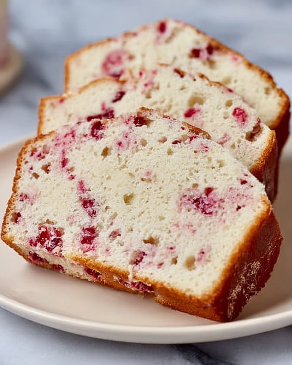 The image shows three thick slices of a soft cake with a light cream color inside, filled with small pieces of red fruit spread throughout. The cake has a slightly golden brown crust on the edges. The slices sit stacked on a white plate with smooth edges, placed on a white marbled background. The texture of the cake looks moist and fluffy, with visible small holes showing its light airiness. Photo taken with an iphone --ar 4:5 --v 7