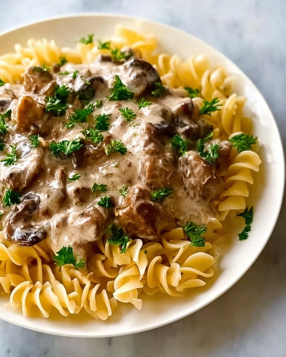 The image shows a close-up of a white plate filled with spiral pasta at the bottom, which is light yellow and slightly shiny. On top of the pasta, there is a creamy sauce layer with pieces of brown mushrooms and chunks of tender-looking meat, all covered in a light beige sauce that looks smooth and thick. Green parsley leaves are sprinkled over the sauce, adding a fresh touch of color. The background is a white marbled surface. Photo taken with an iphone --ar 4:5 --v 7