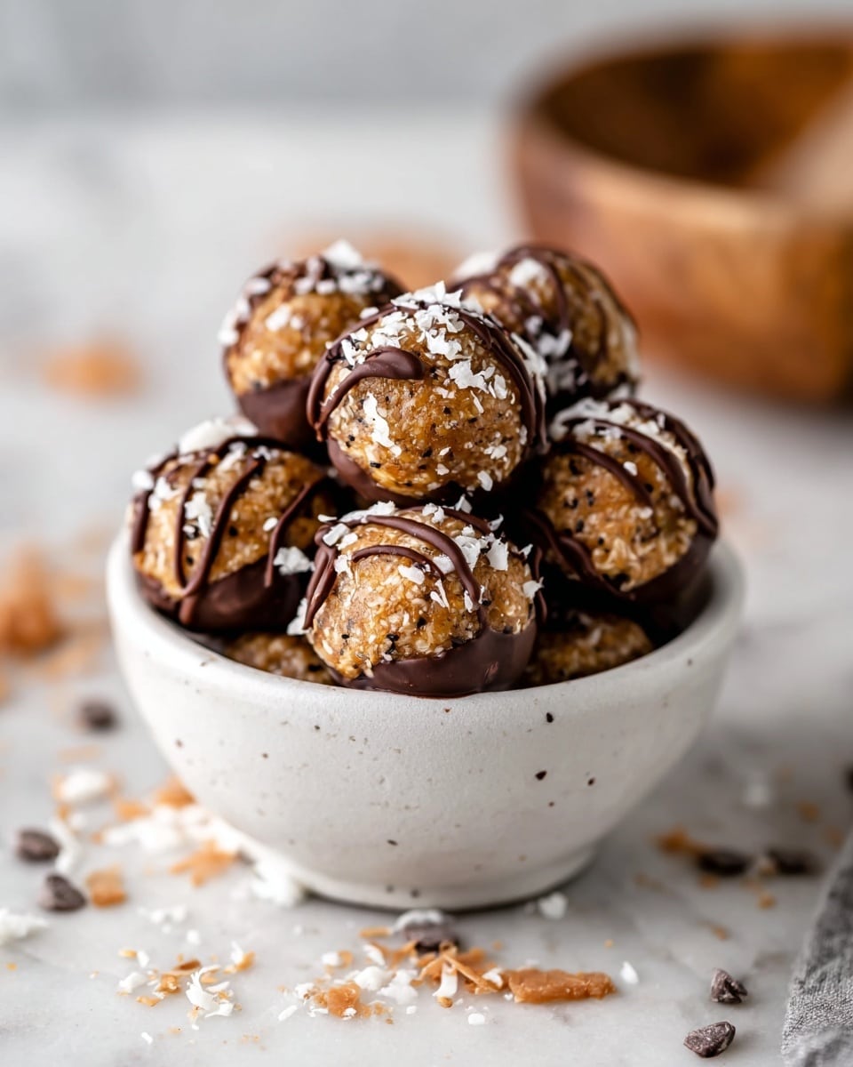 A small white bowl filled with round energy bites, about eight pieces stacked inside. Each bite is light brown with a coarse texture, speckled with tiny dark bits. The bottoms of the bites are dipped in smooth, dark chocolate, while the tops are drizzled with thin lines of the same chocolate and scattered with small white flakes, possibly coconut. The bowl sits on a white marbled surface with some chocolate bits and crumbs around it. In the background, there is a blurred wooden bowl partially visible. photo taken with an iphone --ar 4:5 --v 7