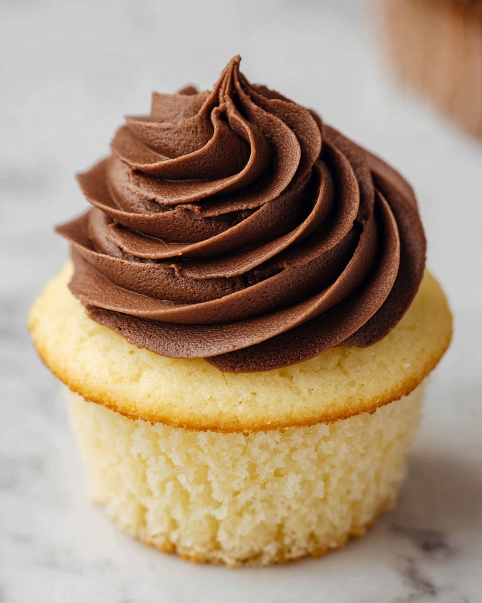 A close-up view of a single cupcake with two distinct layers: a soft, light yellow cake base with a slightly rough texture and a thick swirl of smooth, dark brown chocolate frosting on top. The frosting is piped in a spiral pattern, starting from the outer edge and rising in curls towards the center, creating a peak with defined ridges. The cupcake is set against a white marbled texture background, with a small part of a brown tube showing in the corner. Photo taken with an iphone --ar 4:5 --v 7