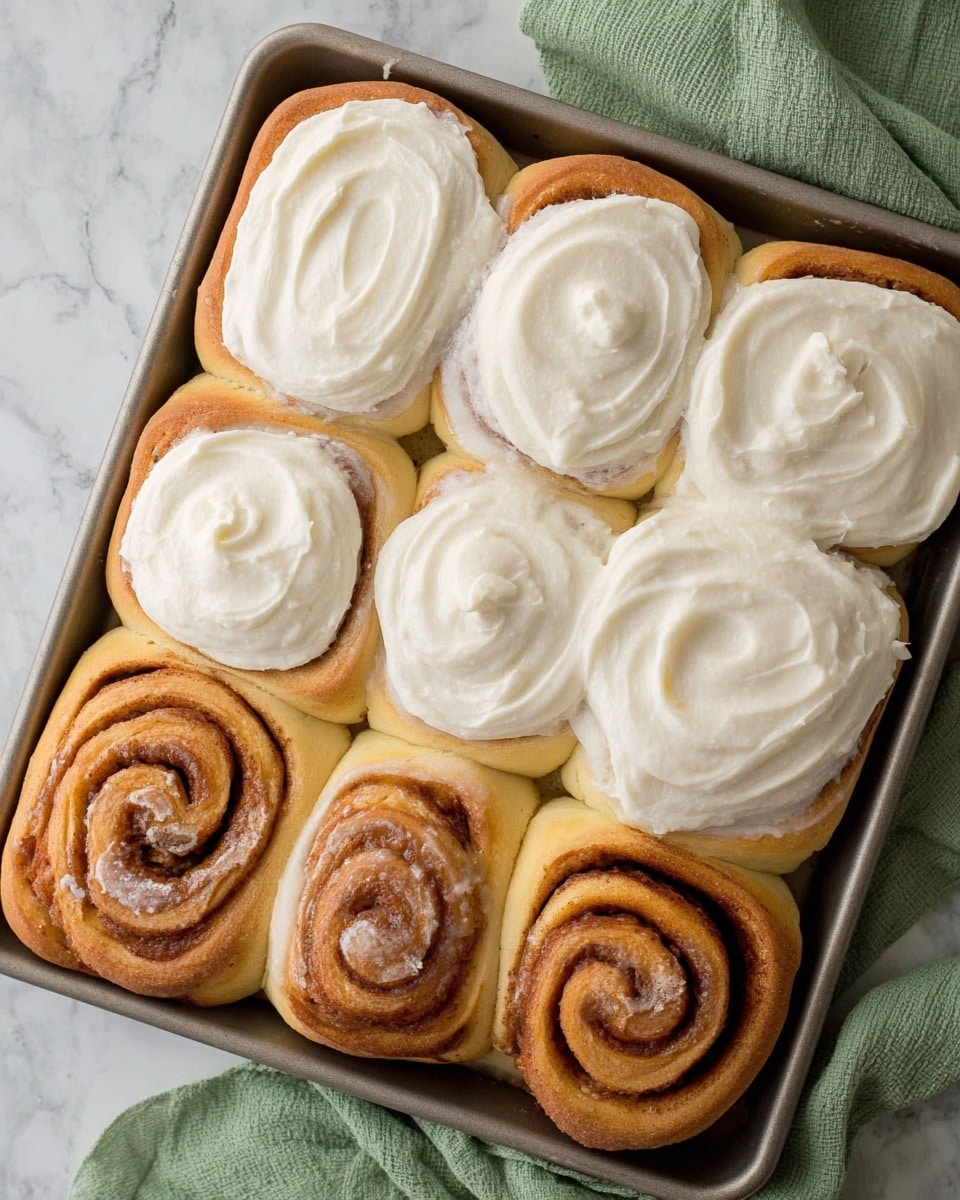 A baking tray filled with nine cinnamon rolls, six of them topped with a thick layer of white creamy frosting that has a smooth, swirled texture; the cinnamon rolls are golden brown with visible spiral layers underneath the frosting. The three unfrosted rolls reveal a shiny, cinnamon-coated swirl pattern with a slightly darker brown color. The tray rests on a white marbled surface, and a green cloth is placed at the top right edge. The photo was taken with an iphone --ar 4:5 --v 7