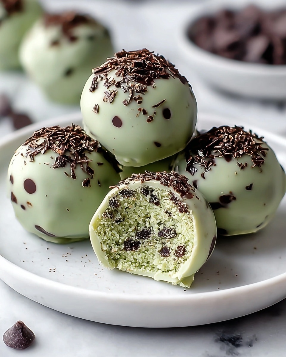 There are five round truffles on a white plate with a white marbled texture beneath. Each truffle has a smooth, pale green outer coating speckled with small dark chocolate bits, and the top is sprinkled with larger chocolate flakes. One truffle is cut open, showing a dense inside that is light green mixed evenly with tiny dark chocolate pieces. Around the plate, a few whole dark chocolate chips are scattered. The photo taken with an iphone --ar 4:5 --v 7
