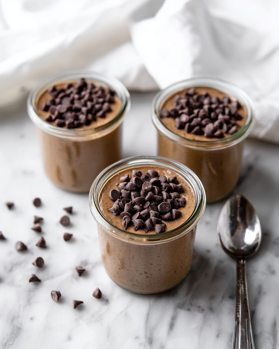 Three clear glass jars are filled with a smooth, light brown creamy dessert. Each jar is topped with a generous layer of dark brown chocolate chips that add texture and richness to the dish. The jars are placed closely together on a white marbled surface, with some chocolate chips scattered around them, adding a casual touch. A shiny silver spoon lies next to the jars on the right side. In the background, a white cloth is softly draped, giving a soft contrast to the sharp details of the jars and the marbled surface. photo taken with an iphone --ar 4:5 --v 7