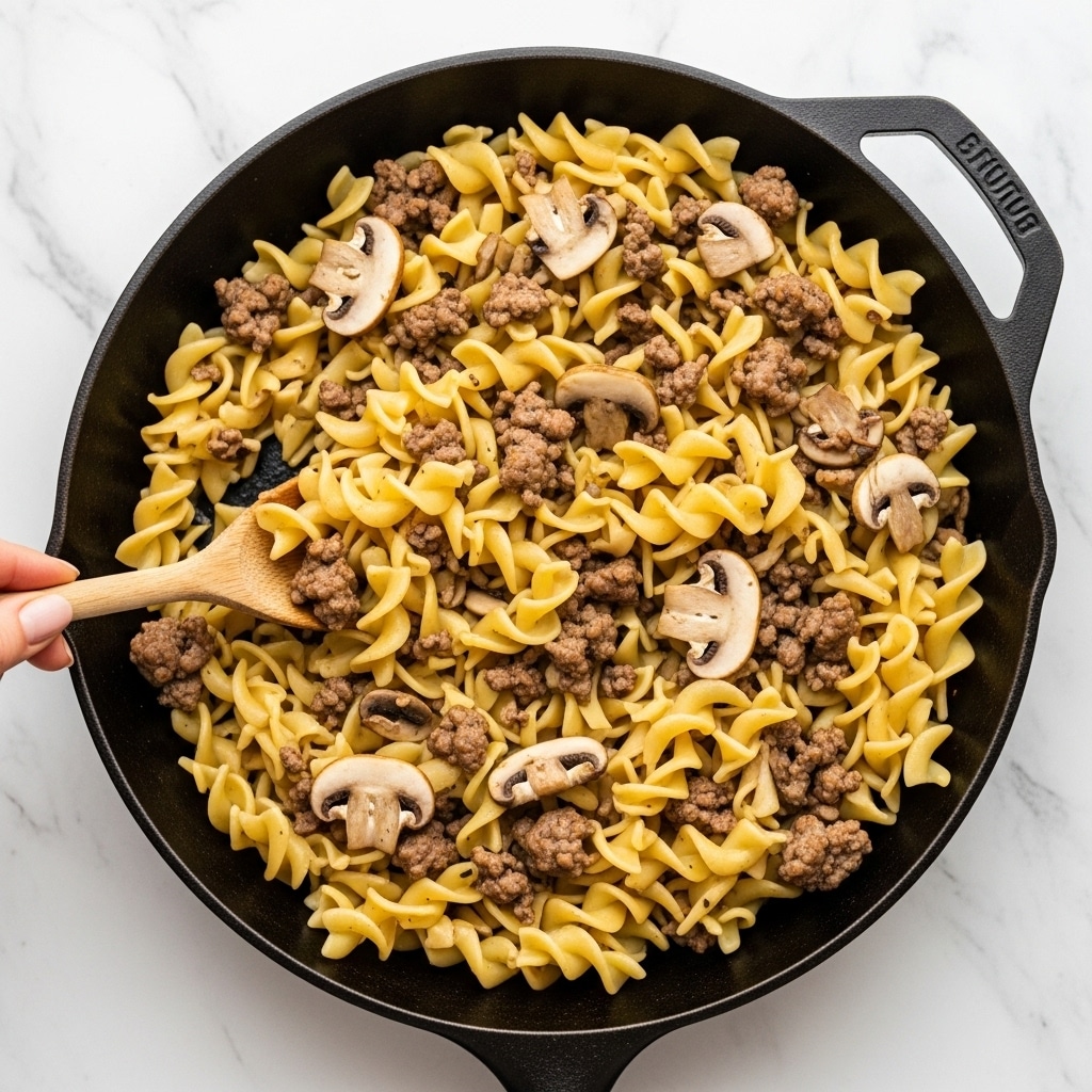 The image shows a black cast iron skillet filled with cooked egg noodles mixed with ground beef and sliced mushrooms. The noodles are light yellow with a soft, slightly ruffled texture, layered evenly throughout the skillet. The ground beef pieces are small, brown, and scattered evenly among the noodles, while the mushrooms are thinly sliced with a light grayish-brown color, blending naturally with the other ingredients. A wooden spoon is partially seen lifting a portion of the mixture near the left side of the skillet, held by a woman's hand. The background is a white marbled surface. Photo taken with an iphone --ar 4:5 --v 7