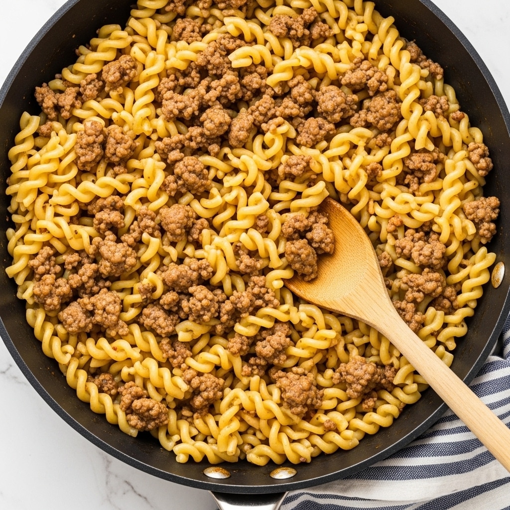 A close-up view of a black pan filled with cooked egg noodles mixed with browned ground meat, showing a layered texture of twisted pasta and small chunks of meat scattered evenly throughout. A wooden spoon is placed inside the pan, mixing the noodles and meat together, with a white marbled surface beneath the pan and a striped blue and white cloth partially visible at the bottom right corner. The colors include light golden noodles, brown meat, and natural wood tone, creating a warm and rustic feel. Photo taken with an iphone --ar 4:5 --v 7