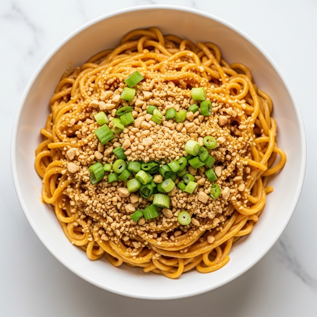 A close-up of creamy noodles placed in a white bowl set on a white marbled surface, with about one main layer of noodles coated in a rich, orange-brown sauce giving them a glossy and smooth texture, topped with a sprinkling of crushed light tan peanuts and bright green chopped scallions scattered evenly across the center, making the dish look fresh and crunchy. Photo taken with an iphone --ar 4:5 --v 7