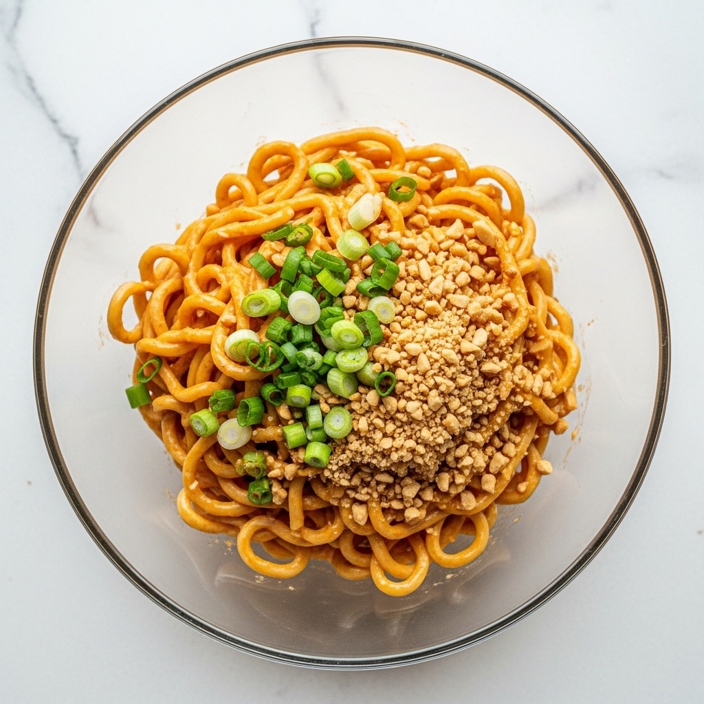 A clear glass bowl filled with thick noodles coated in a creamy orange sauce, topped with chopped green onions and sprinkled with crushed peanuts. The noodles are tangled and glossy, with the green onion pieces and peanut bits creating a colorful contrast on top. The bowl is placed on a white marbled surface. photo taken with an iphone --ar 4:5 --v 7
