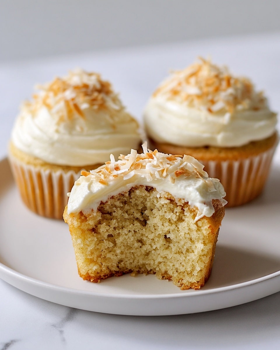 The image shows three light golden cupcakes on a white plate placed on a white marbled surface. The cupcakes have one main layer of soft, moist cake with a rough crumb texture. Each cupcake is topped with a generous swirl of creamy white frosting, smooth and fluffy in texture. On top of the frosting, there is a layer of light brown toasted coconut flakes scattered evenly, adding a crunchy detail. One cupcake is cut in half and placed in front, displaying its airy inside and the thick frosting layer above it. Photo taken with an iphone --ar 4:5 --v 7