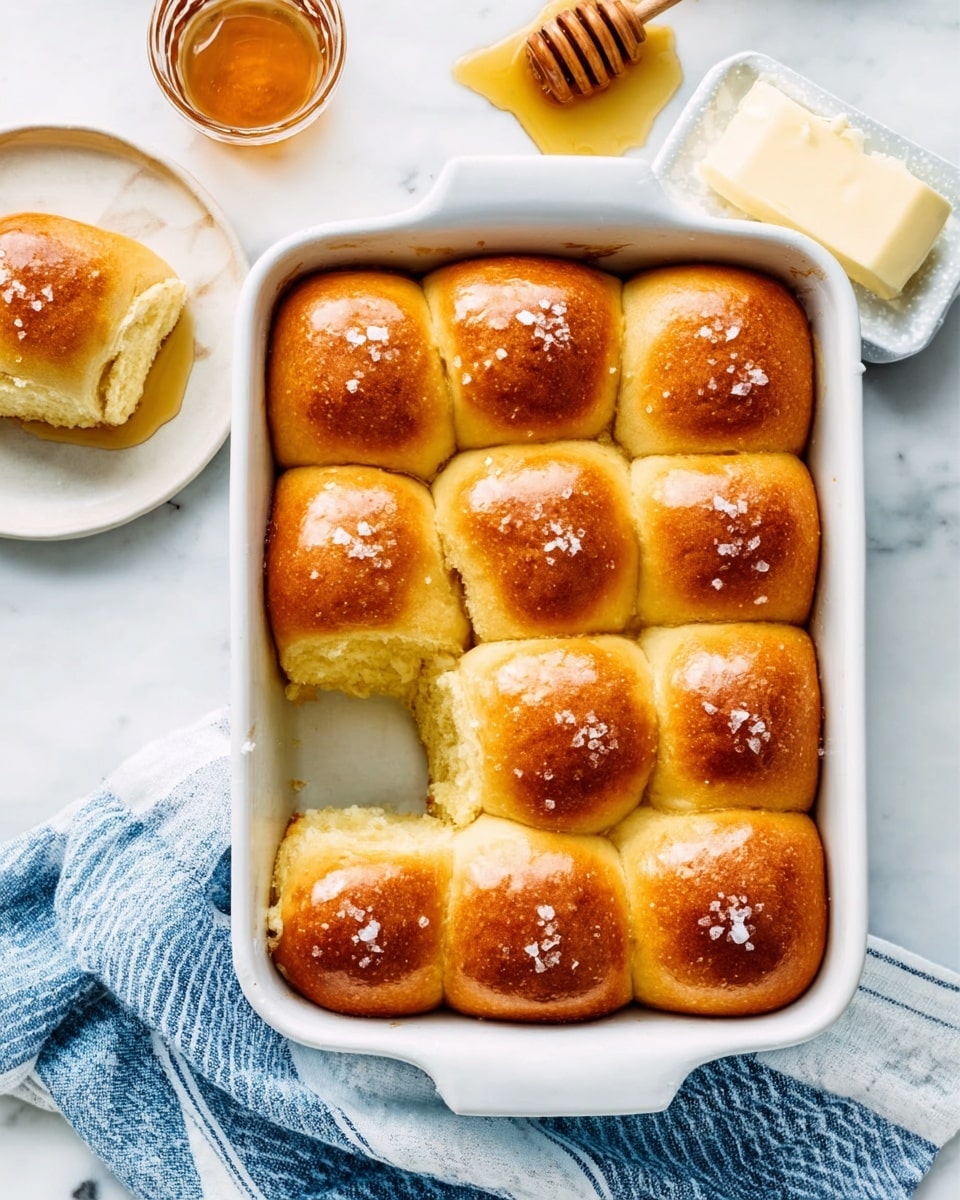 The image shows a white rectangular baking dish filled with twelve golden brown dinner rolls. Each roll has a shiny, slightly glossy top with a few grains of coarse salt sprinkled on some of them. The rolls are tightly packed in three rows and four columns, with a soft and fluffy texture visible where one roll is pulled away, revealing its airy inside. To the upper right side, there is a white square block of butter on a small dish, and above the baking dish, a honey dipper drizzling honey. On the left side, a half-eaten roll sits on a small round white plate, with a blue and white striped kitchen towel placed partially under the dish, all set on a white marbled surface. The photo taken with an iphone --ar 4:5 --v 7