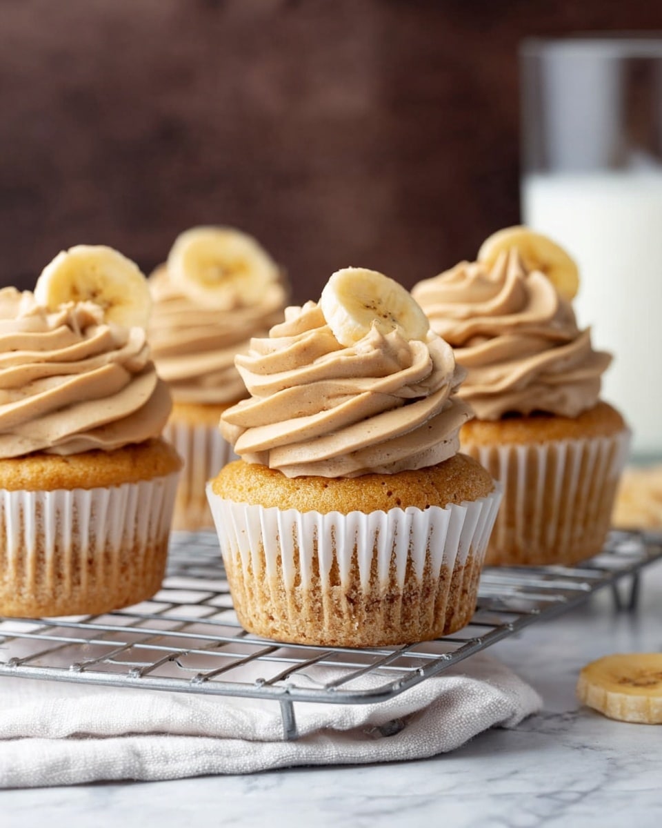 A close-up image of four cupcakes arranged on a silver wire cooling rack over a white cloth, placed on a white marbled textured surface. Each cupcake has two layers: the base is a golden-brown cake in white paper liners with visible crumb texture, and the top is a thick swirl of light brown frosting with a smooth, creamy texture. Each frosting swirl is topped with a single banana chip, which is slightly golden with a crisp texture. The background is blurred with a dark brown color, and there is a glass of milk behind the cupcakes. Photo taken with an iphone --ar 4:5 --v 7