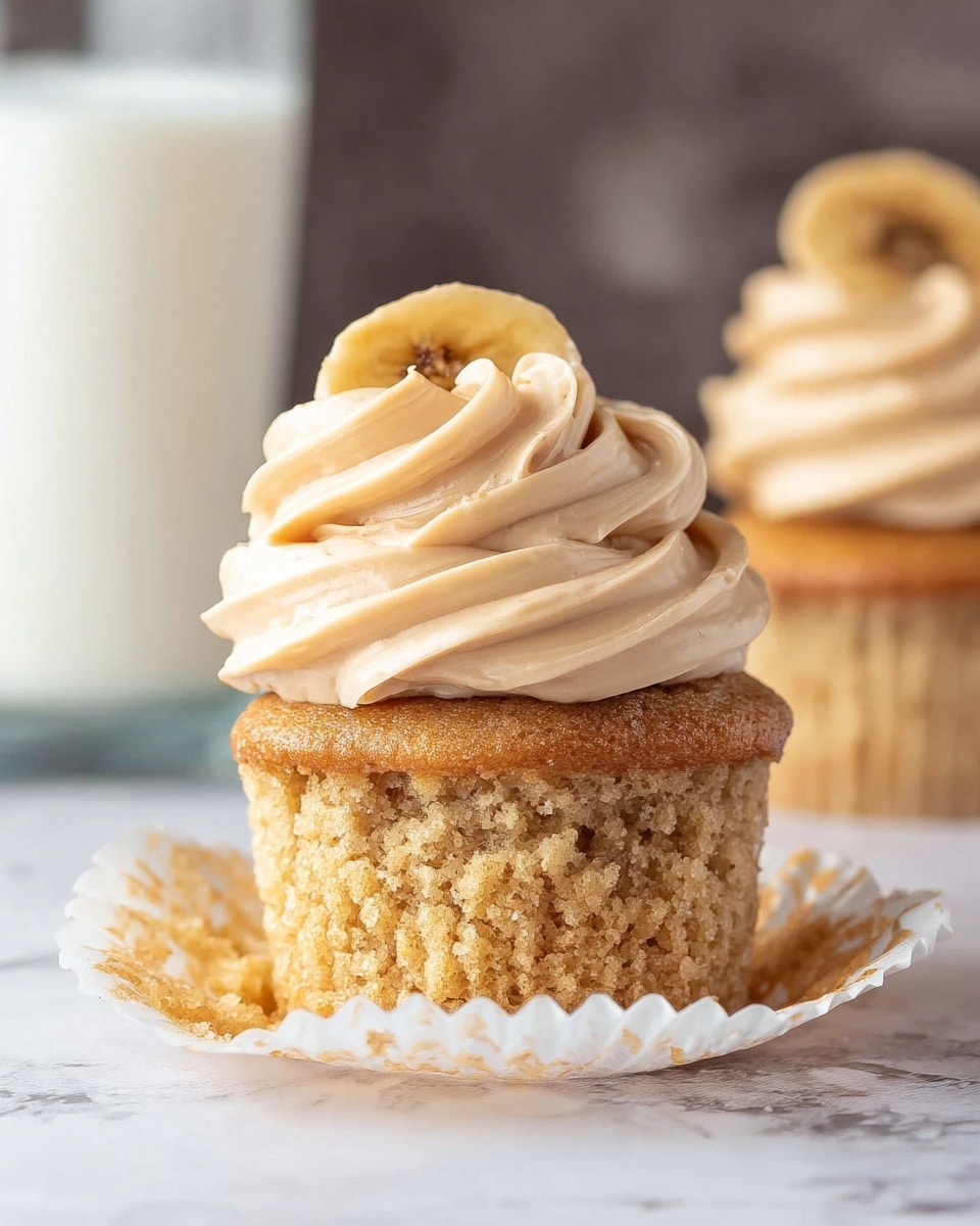 A close-up image of a single cupcake with three layers: the bottom layer is a light golden brown cupcake base with a soft, crumbly texture, partially wrapped in a slightly opened white paper liner; the middle layer is a large swirl of creamy, light tan frosting with a smooth and fluffy texture sitting on top of the cupcake; the top layer is a single dried banana chip placed upright in the frosting. In the blurry background, there is another cupcake with similar frosting and a glass of white milk. The surface is a white marbled texture. photo taken with an iphone --ar 4:5 --v 7