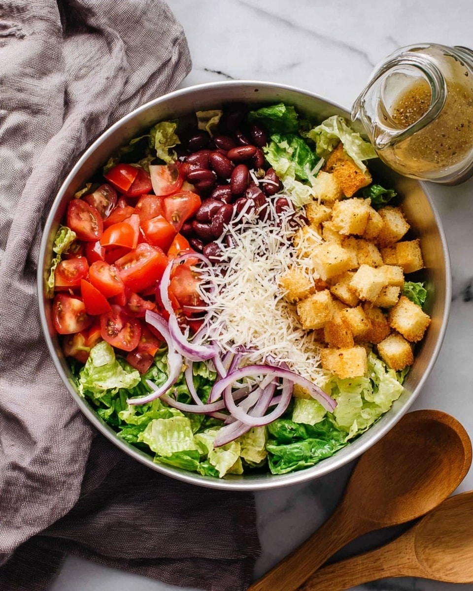 The image shows a white metal bowl filled with fresh salad divided into neat sections. The bottom layer is crisp green lettuce leaves, followed by bright red tomato chunks on the left side. Above the tomatoes are purple kidney beans, some thin purple onion slices are scattered. To the right, golden-brown crouton cubes are sprinkled with white grated cheese. A small glass jar of salad dressing leans against the bowl's edge. The bowl sits on a white marbled surface with a soft gray cloth in the background and two wooden salad spoons partly visible at the bottom. photo taken with an iphone --ar 4:5 --v 7