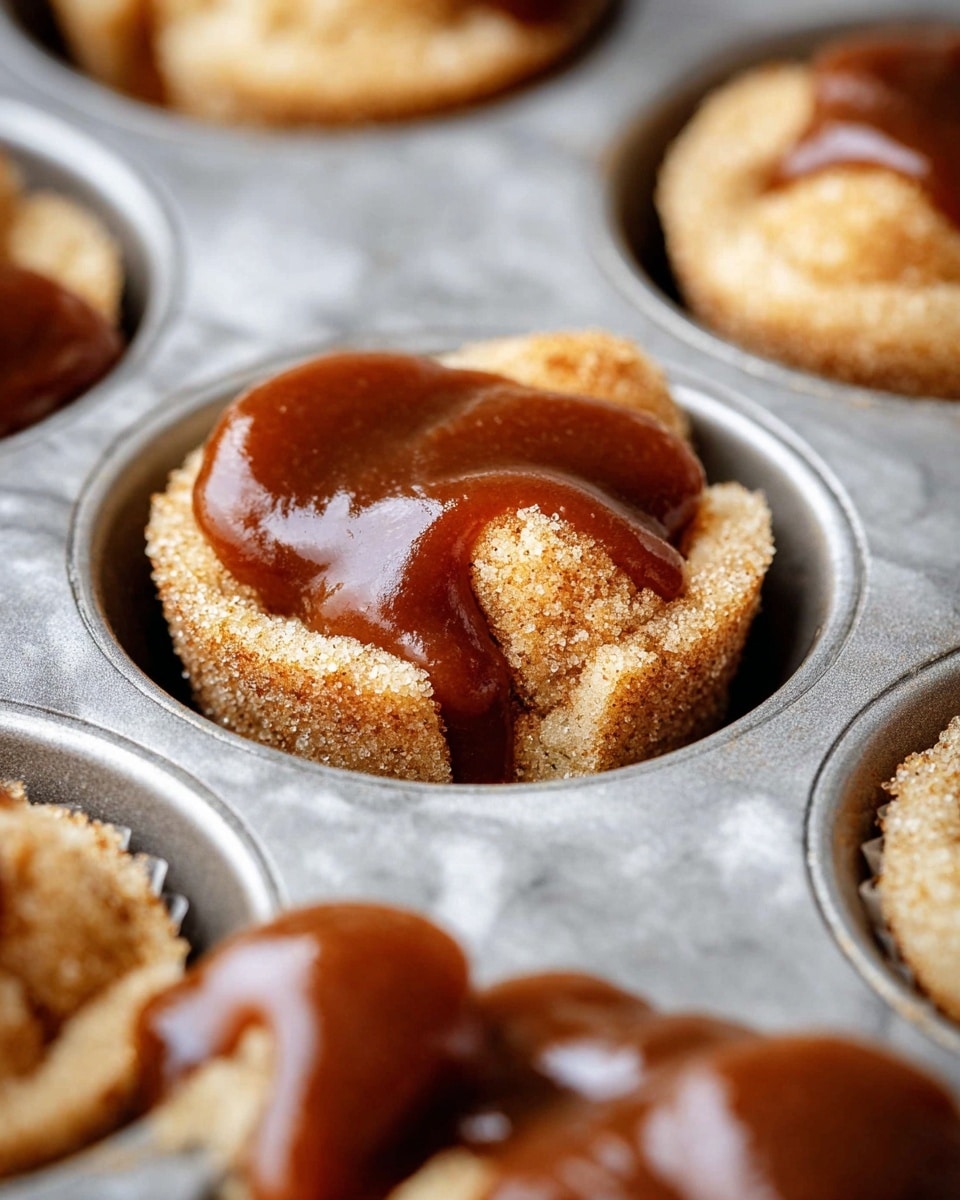 The image shows a close-up of a metal muffin tray filled with bite-sized dough pieces covered in cinnamon sugar on the bottom layer, topped with a thick, glossy brown sauce that looks smooth and sticky, filling the muffin cup almost to the top, with some sauce slightly spilling over the dough pieces. The dough pieces are light golden brown with a coarse, grainy cinnamon texture. The background is a white marbled surface. photo taken with an iphone --ar 4:5 --v 7