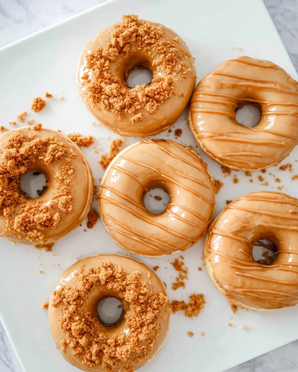 The image shows six round donuts on a white plate, placed on a white marbled surface. Each donut is coated with a smooth light brown caramel glaze that covers the top and partly drips down the sides. Three donuts have a crumbly, golden brown cookie topping scattered unevenly on the glaze. Two donuts are decorated with thin caramel drizzle lines crossing over the glaze. One donut on the top right only has the plain caramel glaze without any topping or drizzle. Some crumbs are scattered around the donuts on the plate, adding texture to the scene. photo taken with an iphone --ar 4:5 --v 7
