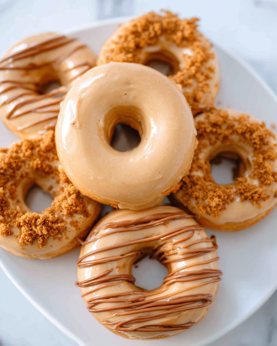 A white plate holds six round donuts with a hole in the middle, arranged with one donut in the center on top of five others. The center donut is coated with a smooth, shiny light brown glaze. The five donuts underneath have two different toppings: three have the same light brown glaze base with crunchy brown crumbs sprinkled on half of their tops, and two are decorated with thin drizzle lines of darker brown frosting over a light brown glazed surface. The white marbled texture background shows softly behind the plate. photo taken with an iphone --ar 4:5 --v 7