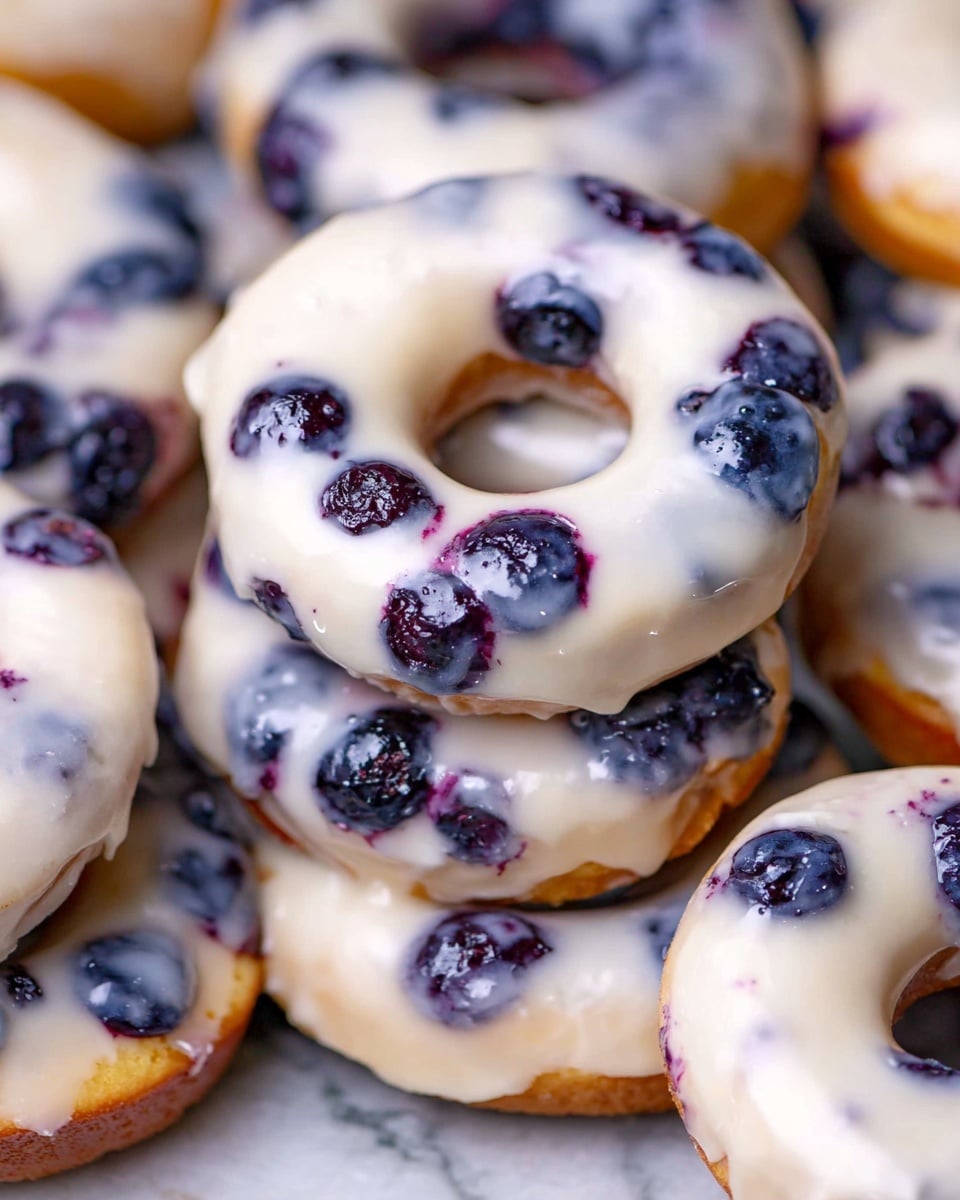 A close-up view of several blueberry donuts stacked and overlapping each other on a white marbled surface; each donut has a light golden base clearly studded with whole dark blueberries with deep purple spots where berries burst, all topped with a smooth, glossy white glaze that evenly coats the surface with slight raised texture, highlighting the berries beneath the glaze and creating a soft, shiny finish across the tops. Photo taken with an iphone --ar 4:5 --v 7