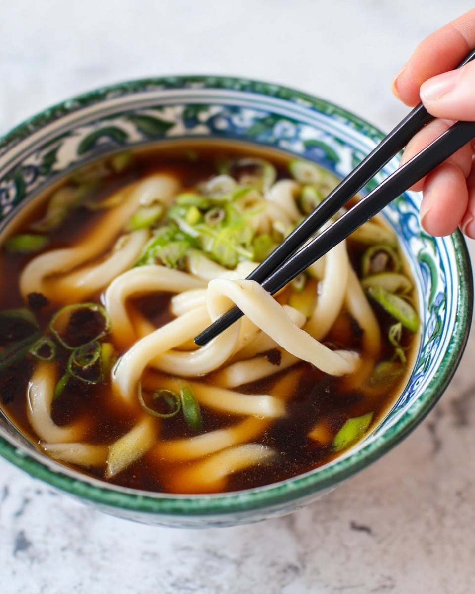 A close-up view of a white bowl with green and blue patterns around the rim, filled with dark brown broth. Thick, creamy white udon noodles float in the broth, some held delicately by a woman's hand using black chopsticks. Small green chopped scallions are scattered on the surface, adding a fresh pop of color. The bowl sits on a white marbled surface, creating a clean and bright setting. photo taken with an iphone --ar 4:5 --v 7